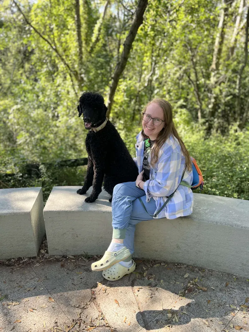 Hannah Forstall sitting on a stone bench in a sunny park with her black standard poodle Siegfried